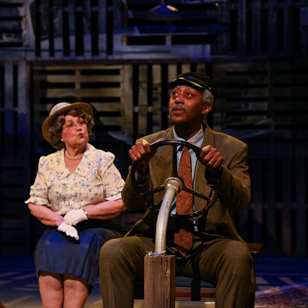 Man acting like he's driving with a steering wheel on a chair talking to older woman.