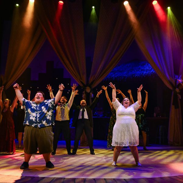 Ensemble cast singing on stage in Hawaiian shirts with hand in air.