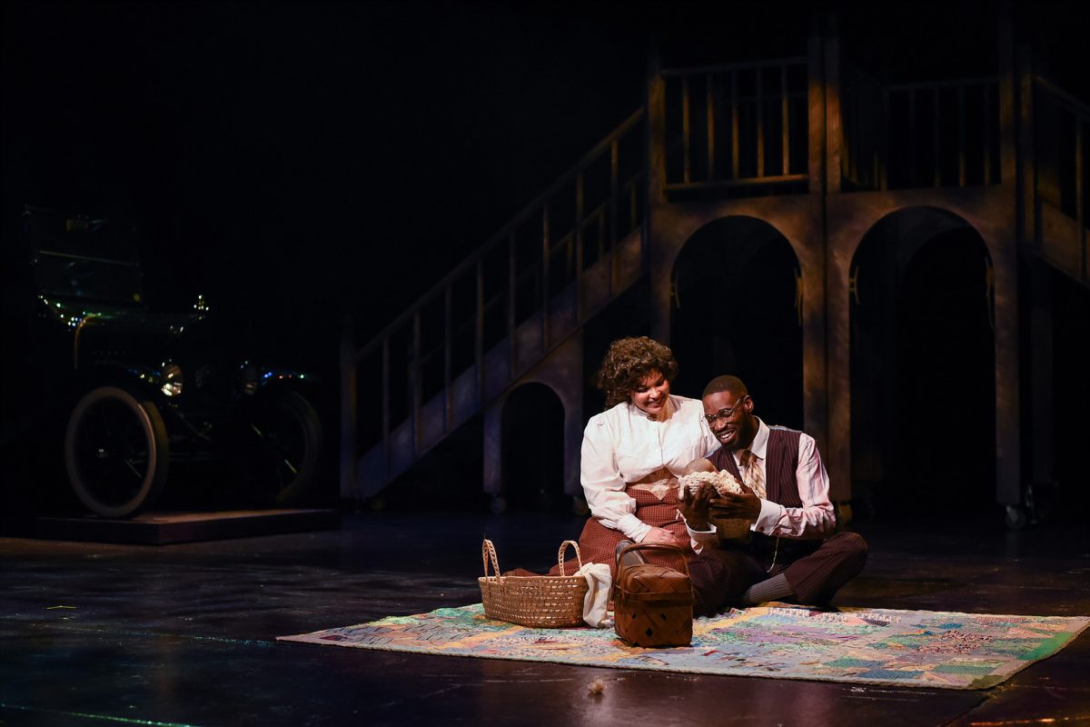 Man and woman on stage, having a picnic, and holding a newborn baby.